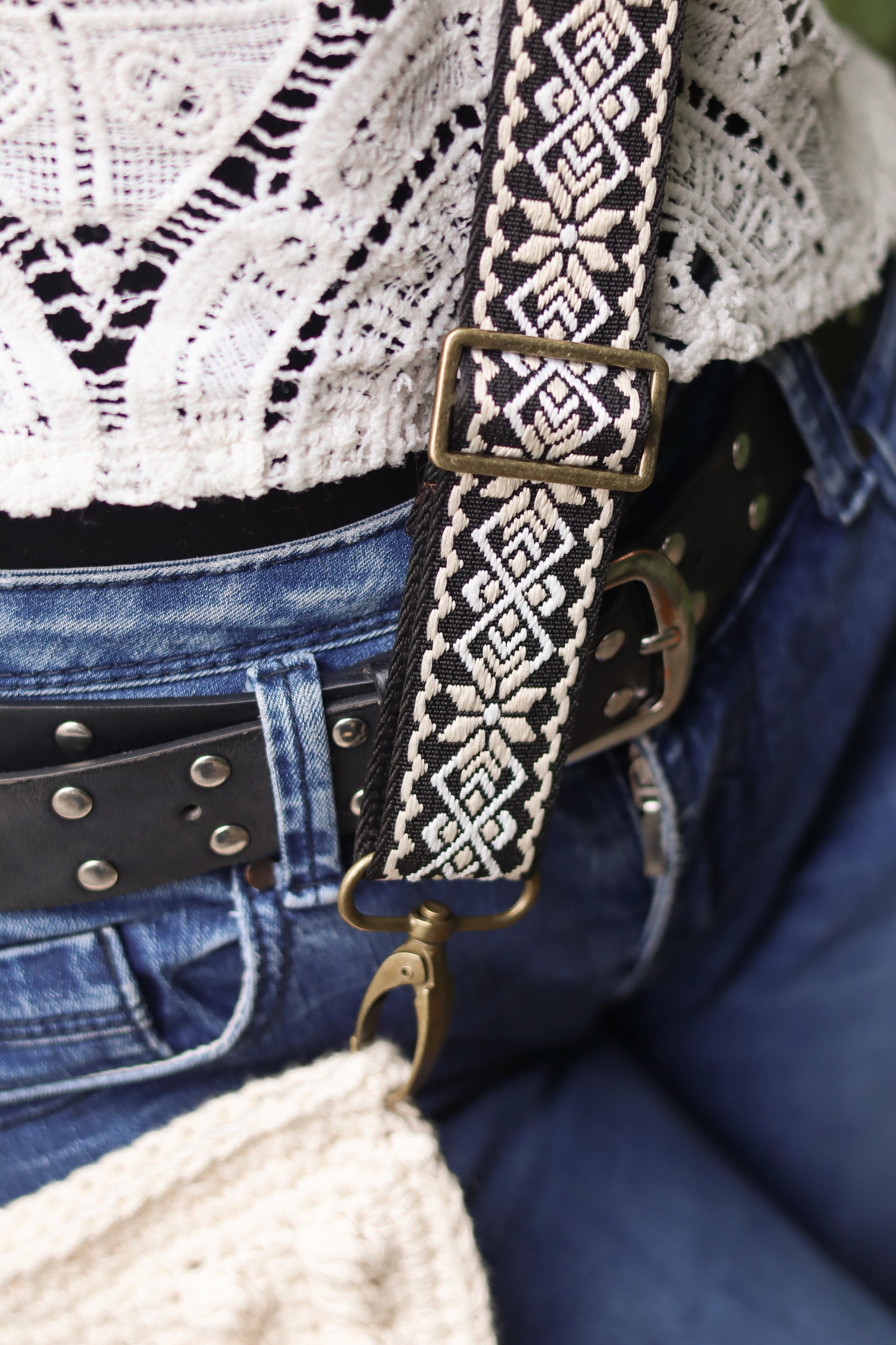 Close-up of a woven boho strap with a crochet handbag on a person wearing a white lace top and blue jeans.