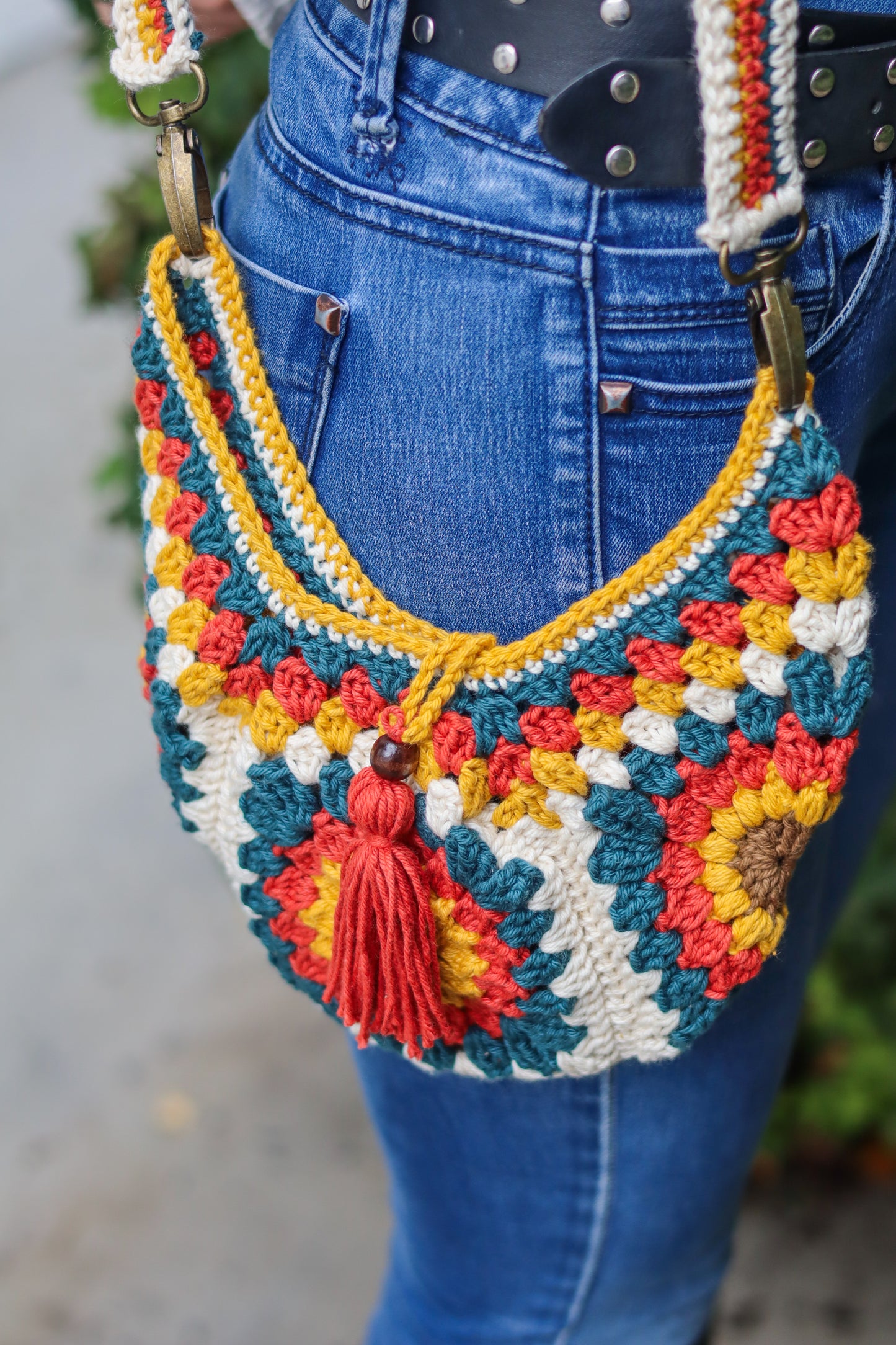 Colorful crocheted handbag with floral patterns and a tassel, worn over blue jeans.